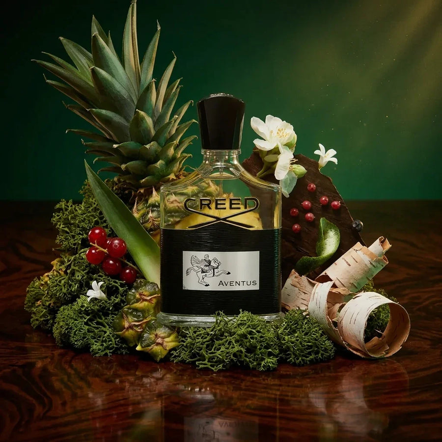 Creed Aventus perfume bottle surrounded by pineapple, flowers, berries, moss, and birch bark