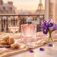 Luxury perfume bottle with pink liquid, macarons, champagne, and purple flowers on table.